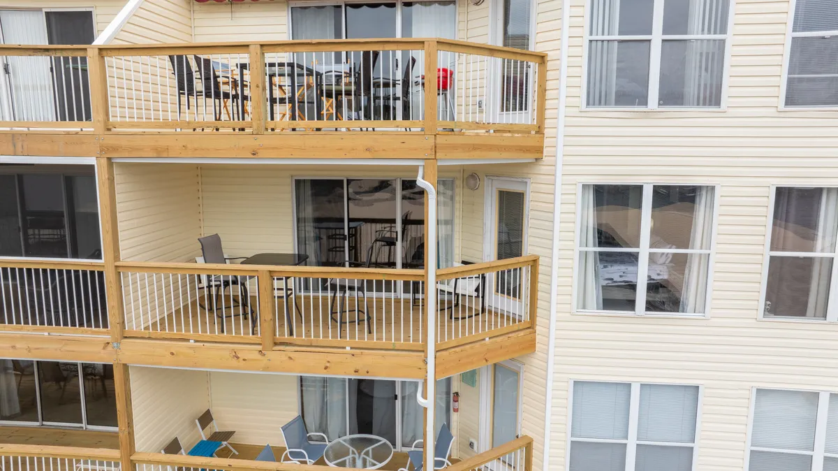 The photo features a cozy balcony equipped with chairs and a table, framed by large windows and a wooden railing. The outdoor setup invites quiet conversation, shared meals, or simply soaking in the natural surroundings at sunrise or twilight.