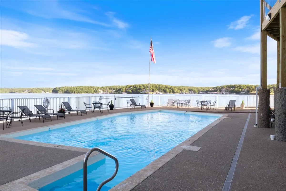 A sparkling swimming pool sits just above the lake, complete with deck chairs for lounging and an American flag waving in the background. The elevated position provides panoramic lake views, while guests enjoy swimming, sunbathing, and socializing in a peaceful, scenic setting.