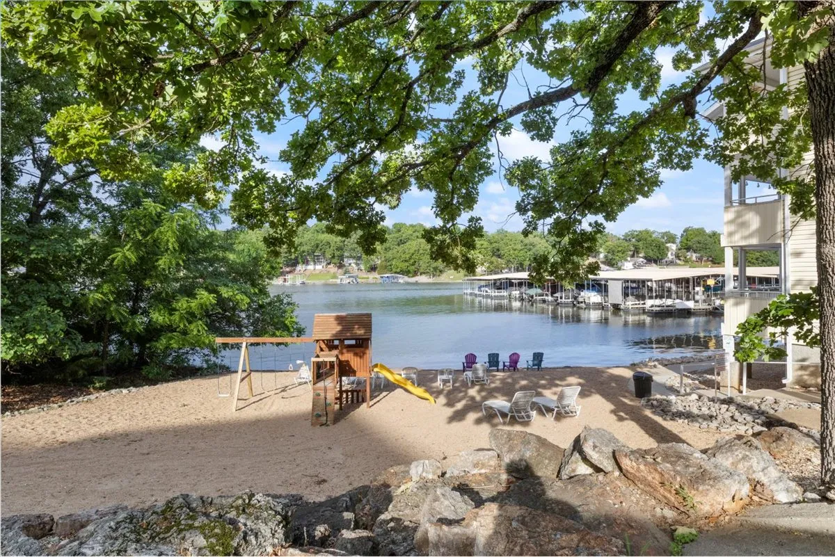 A waterfront beach area featuring a vibrant playground structure, chairs, and trees. The scene is full of activity, with space for swimming, playing, relaxing, and socializing, set against the peaceful backdrop of the lake.