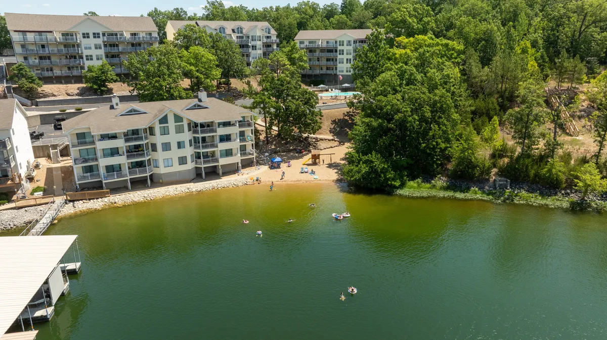 A sweeping view of the lakefront beach area, with families sunbathing, swimming, and enjoying water activities. The residential buildings and green trees frame the scene, creating a carefree, leisurely atmosphere ideal for making summer memories.