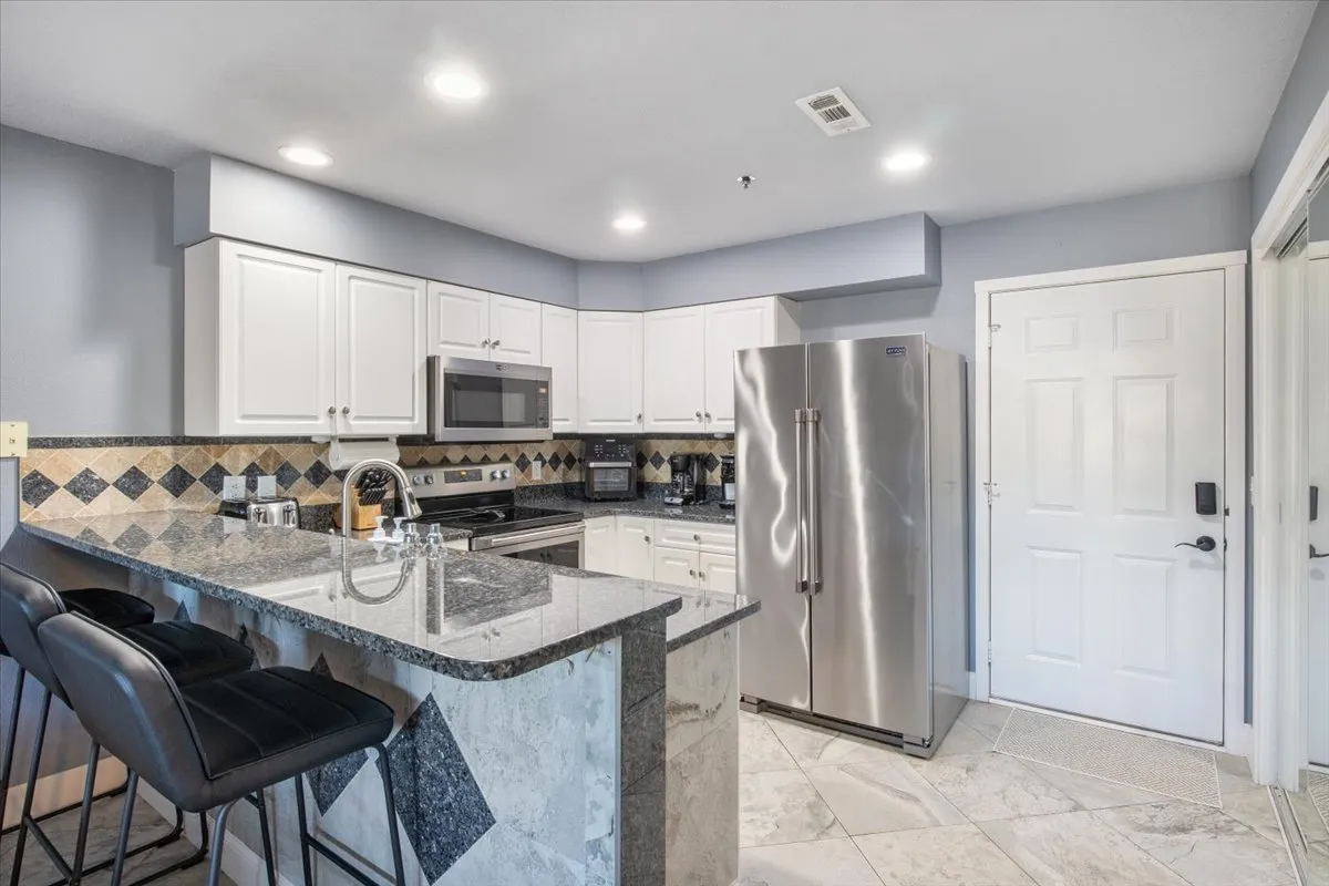 A welcoming kitchen scene with granite countertops, sleek stainless steel appliances, stylish bar stools at the counter, and a decorative tile backsplash. The space is designed for both cooking and socializing, making it easy to engage while preparing meals.