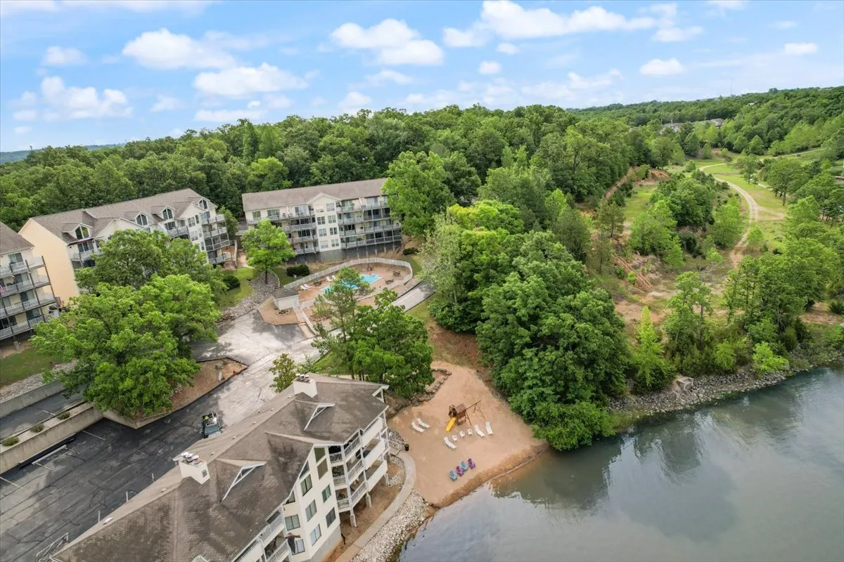 This exterior image captures the lakefront community with visible pools, lush greenery, residential buildings, a clear play area, and a pathway leading toward the waterfront. Families are seen enjoying various activities such as swimming, walking, picnicking, and playing on the sandy beach, all set against a tranquil and inviting lakeside backdrop.