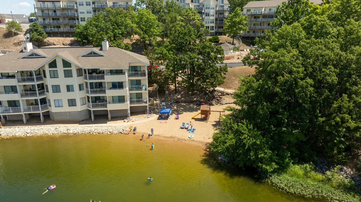 A lively lakefront beach scene with a sandy area, trees, building balconies, and people enjoying various activities like swimming, sunbathing, and playing in the sand. The mood is joyful and carefree, capturing the essence of lakeside leisure and family-friendly fun.