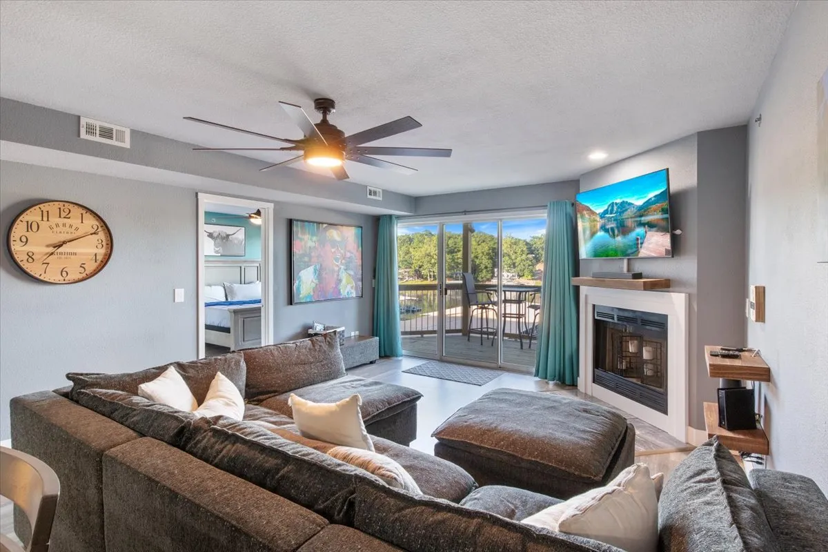 A spacious living area with a large sectional sofa facing a flat-screen television and a modern fireplace, flanked by floor-to-ceiling windows that reveal sweeping views outside. The setup suggests a space designed for movie nights, gatherings, and relaxation while enjoying natural light and the view.